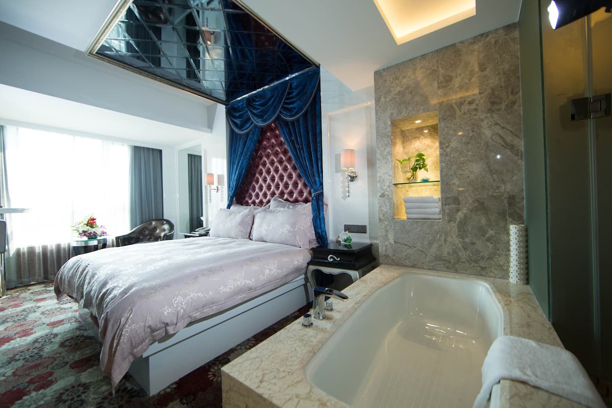Una habitación de hotel moderna con una cama grande, una zona de estar separada, un baño con bañera y un ventanal con cortinas.
