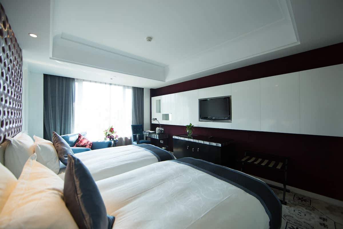 Habitación de hotel con dos camas, un escritorio con silla, un televisor de pantalla plana en la pared y una ventana con cortinas.