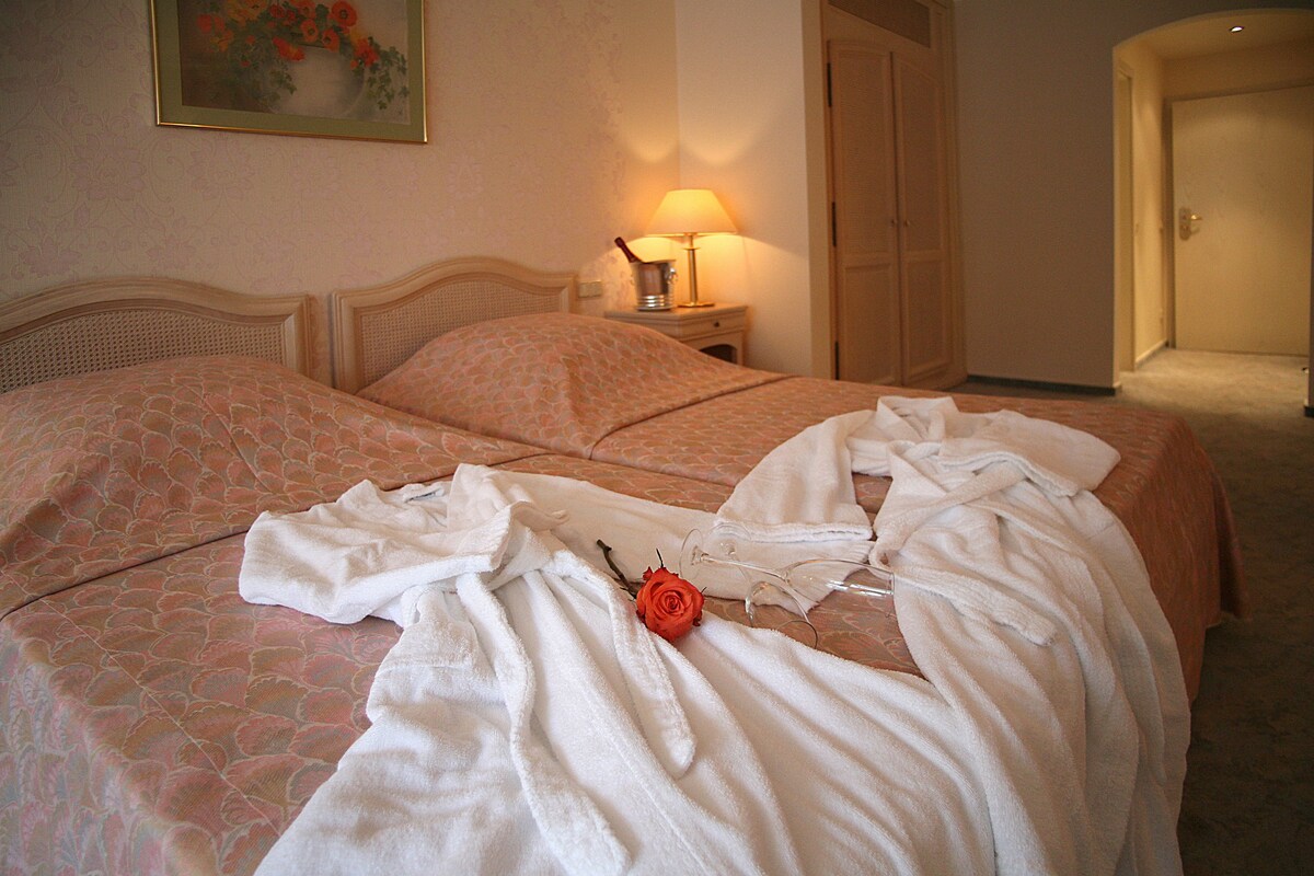 A hotel room with a double bed, a bedside table with a lamp, and a door leading to another room.