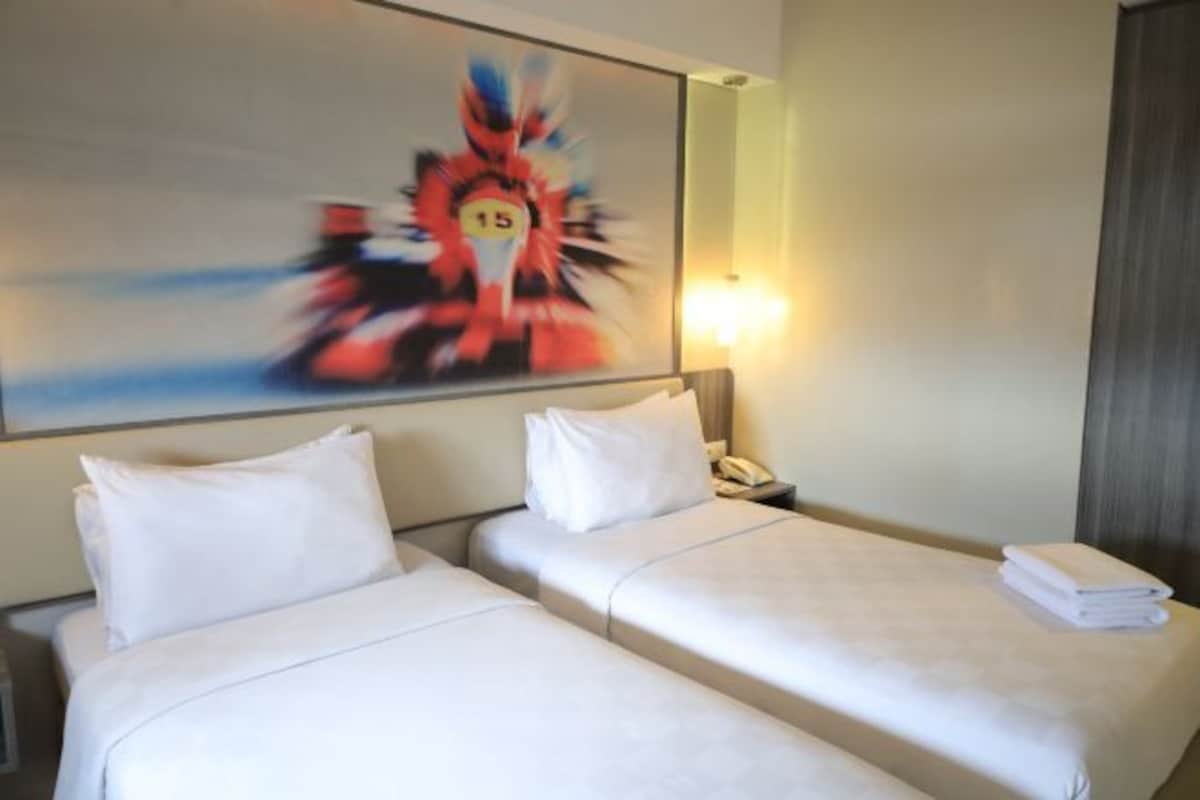 A hotel room with two beds, a painting on the wall, a bedside lamp, and a folded towel on the right bed.