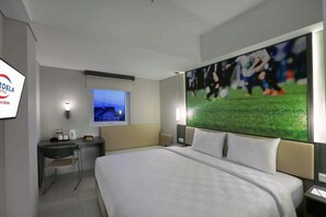 Habitación de hotel con una cama grande, un escritorio con silla, una ventana con vistas y un televisor de pared mostrando un partido deportivo.