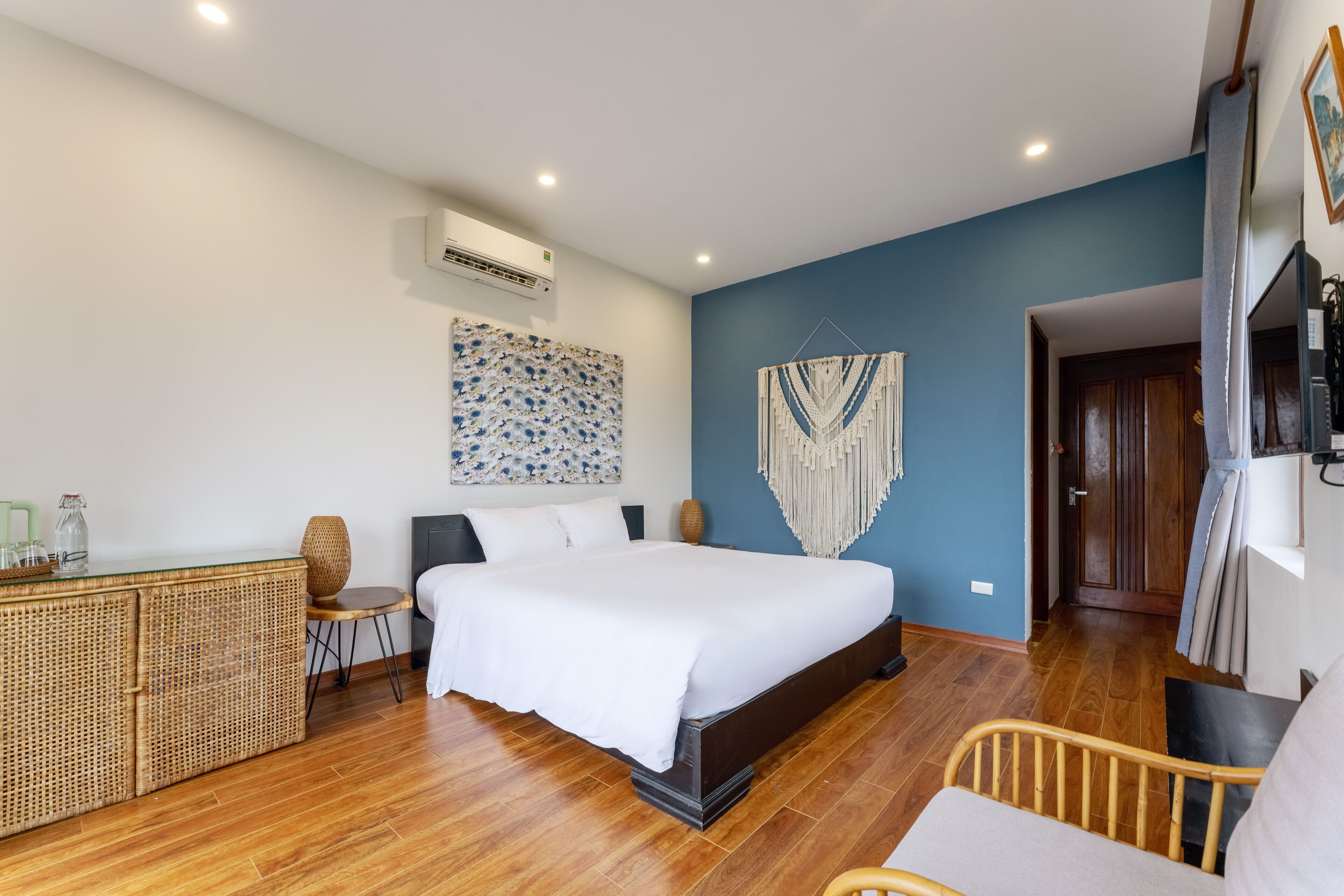 A bedroom with a large bed, a bedside table, a wicker cabinet, a chair, and a decorative wall hanging.
