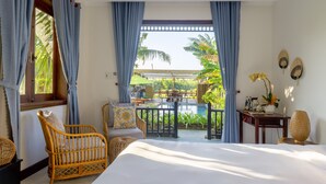 A bedroom with a large bed, a wicker chair, a small table with a vase, and a view of an outdoor pool area. - New Sunshine Homestay (Da Nang)