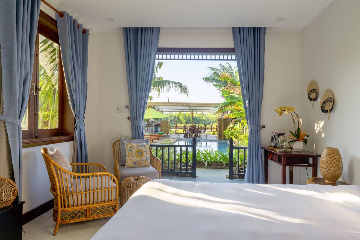 A bedroom with a large bed, a wicker chair, a small table with a vase, and a view of an outdoor pool area.