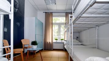 A dormitory room with bunk beds, a small table with chairs, a potted plant, and a window with curtains.