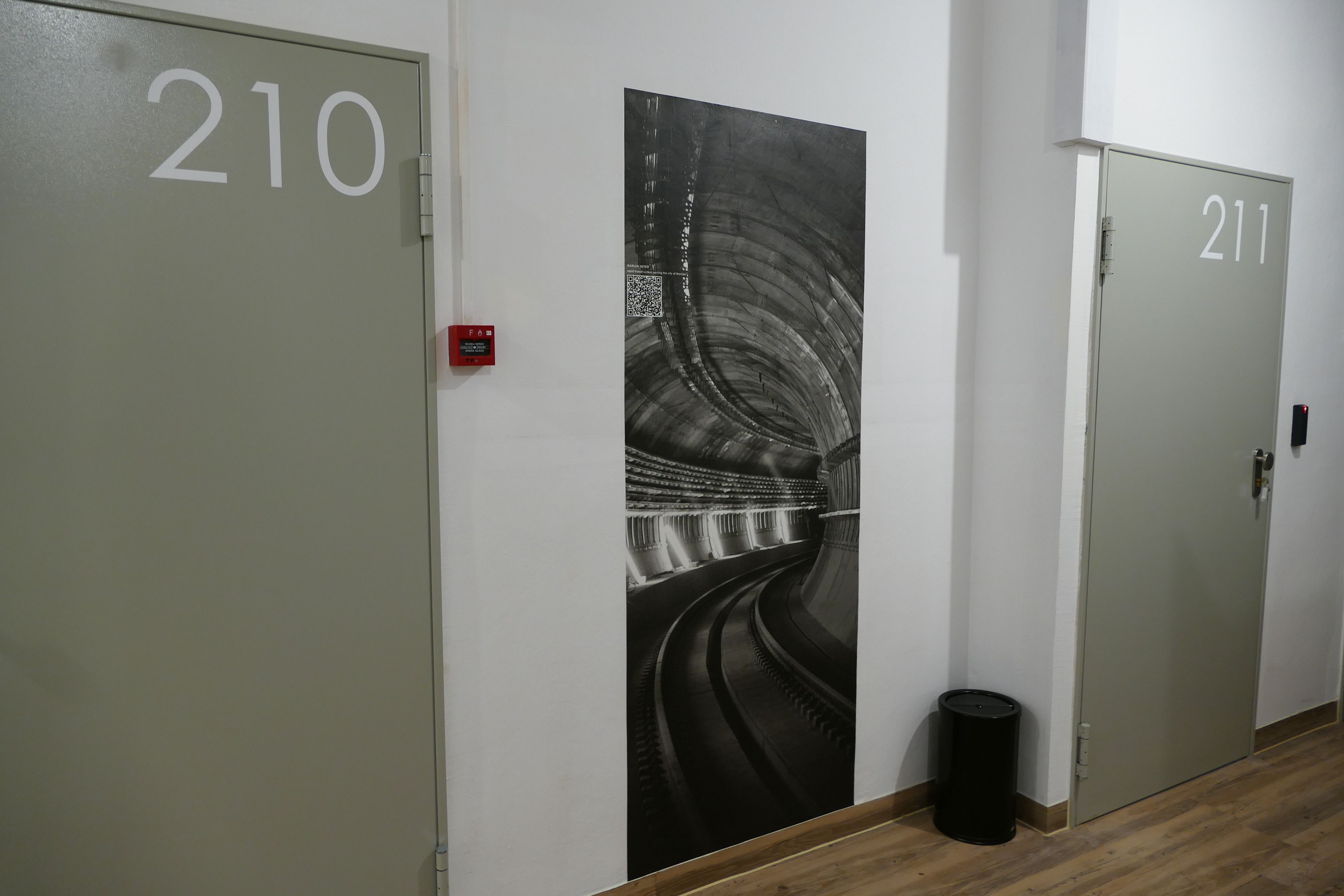 A hallway with doors numbered 210 and 211, a black and white mural of a tunnel, and a trash can.