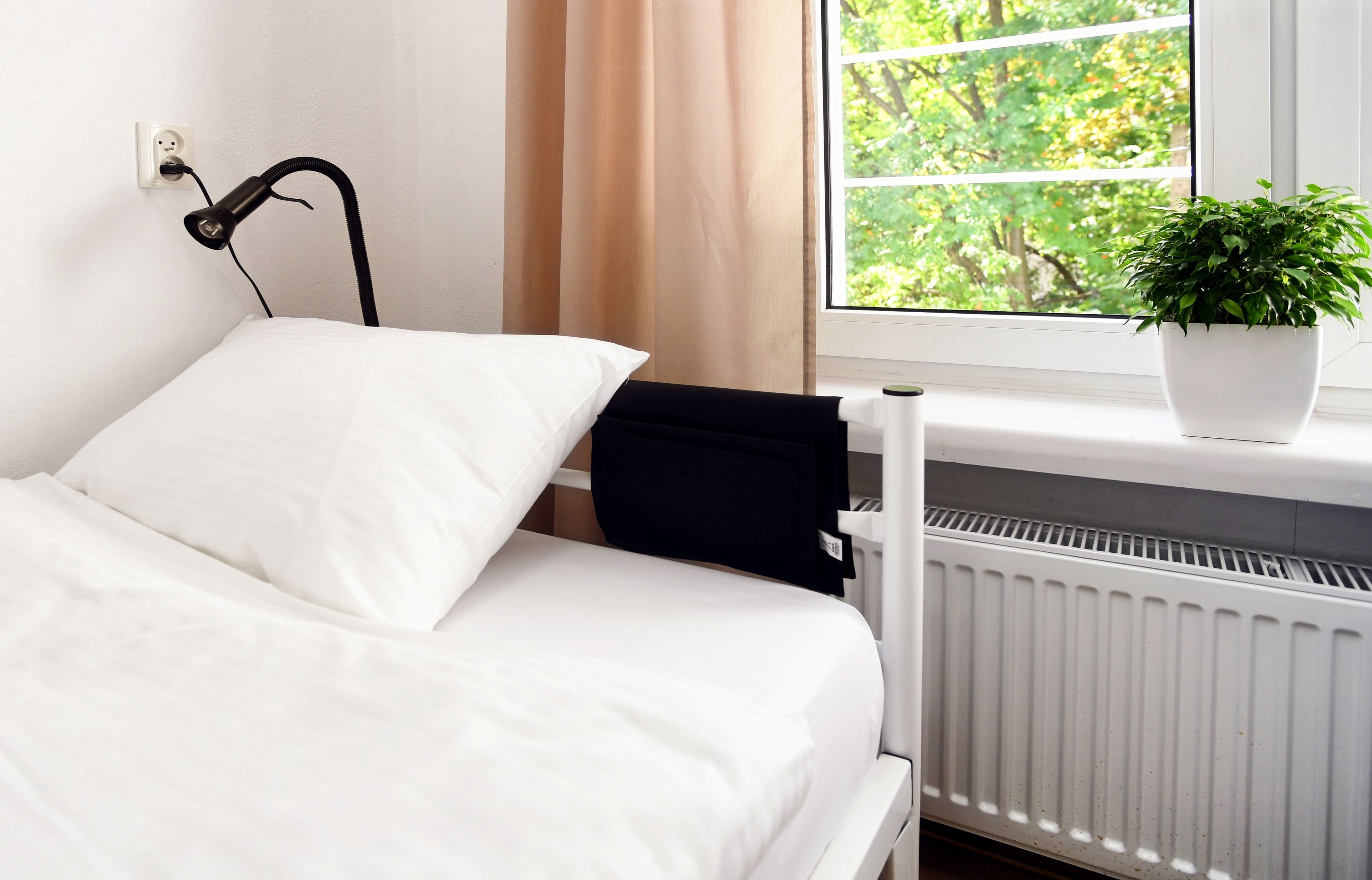 Een netjes opgemaakt bed met witte beddengoed, een zwarte stoel, een plant in een pot op een vensterbank en een radiator onder het raam.