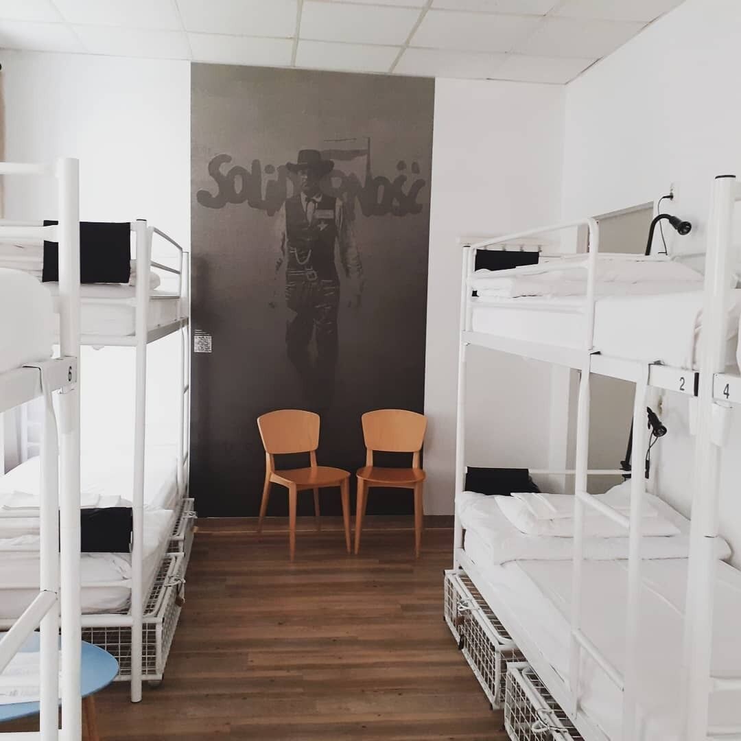A dormitory room with bunk beds, wooden chairs, and a wall with a mural.