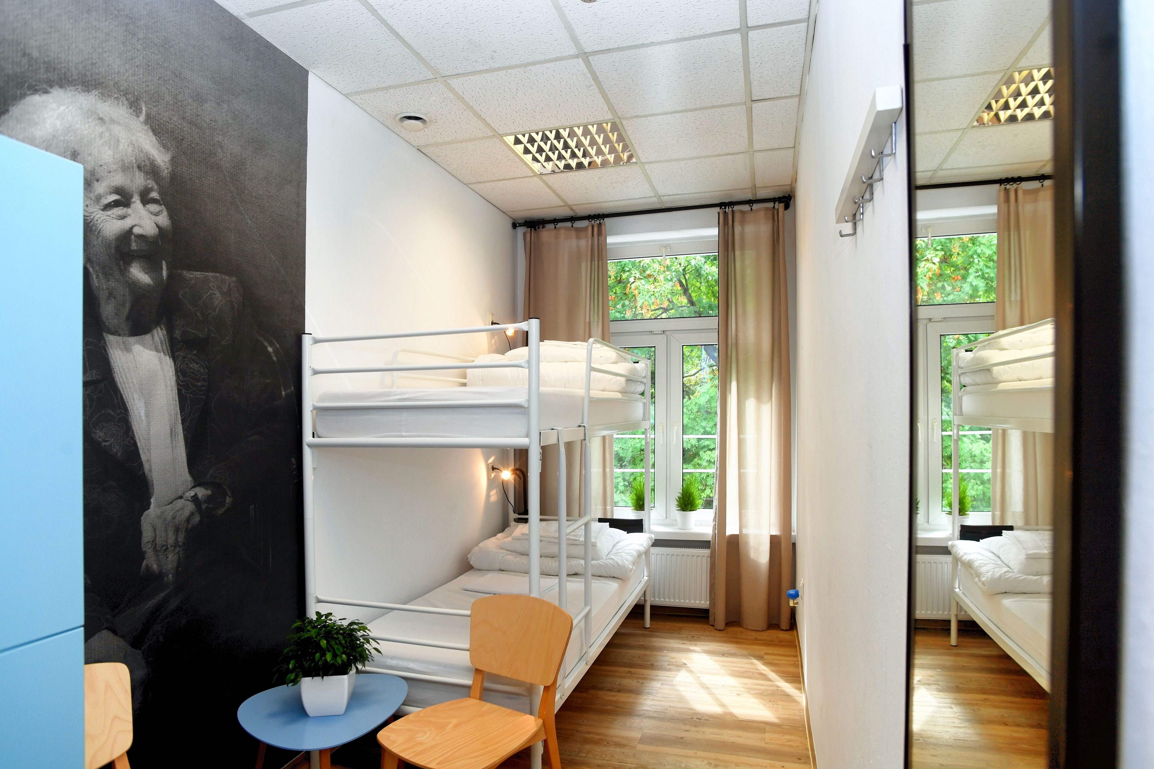 Um quarto de estudante com beliches, uma pequena mesa com uma planta e um mural na parede.