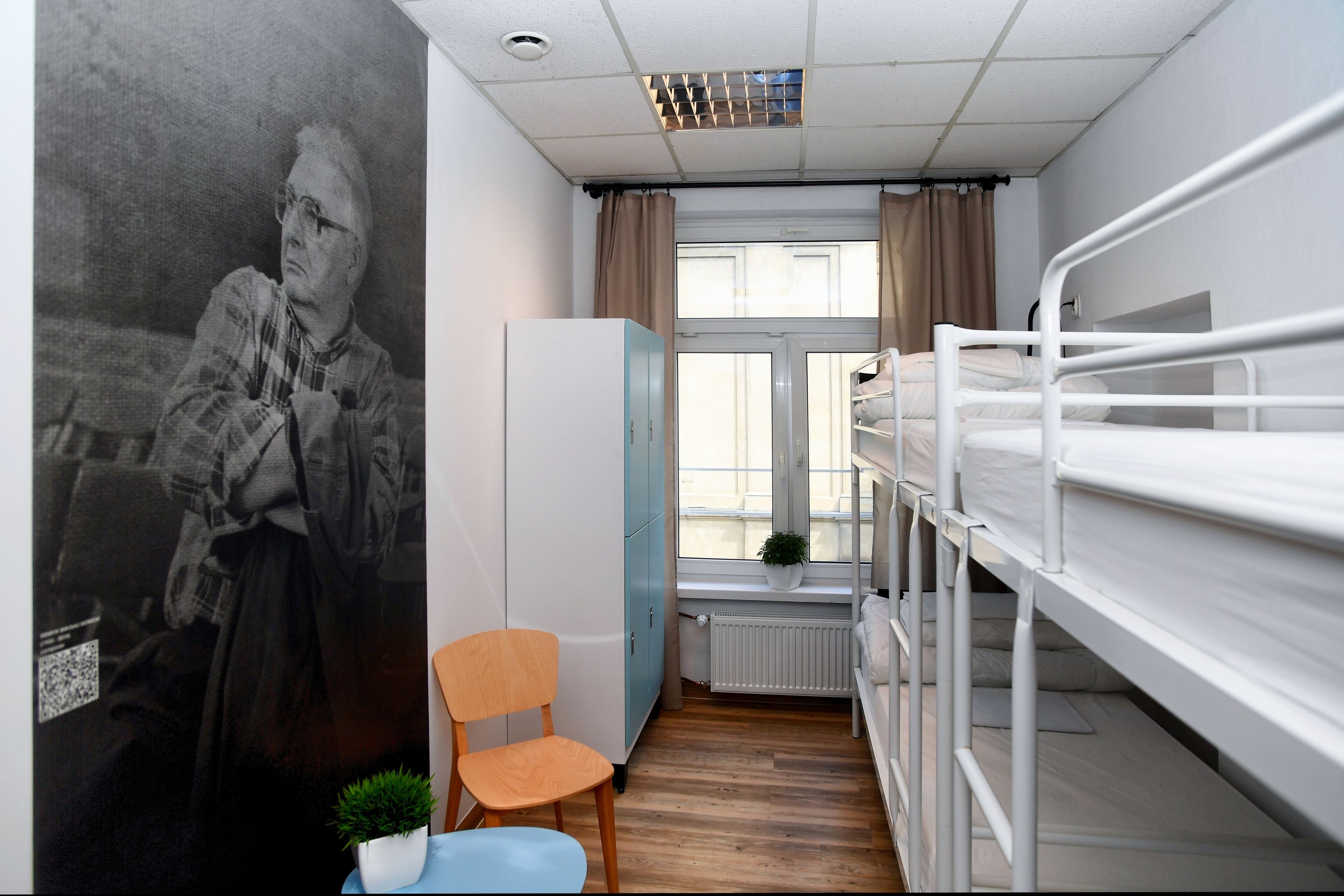 A dormitory room with bunk beds, a wardrobe, a chair, a radiator, and a window with curtains.