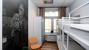 A dormitory room with bunk beds, a wardrobe, a chair, a radiator, and a window with curtains.