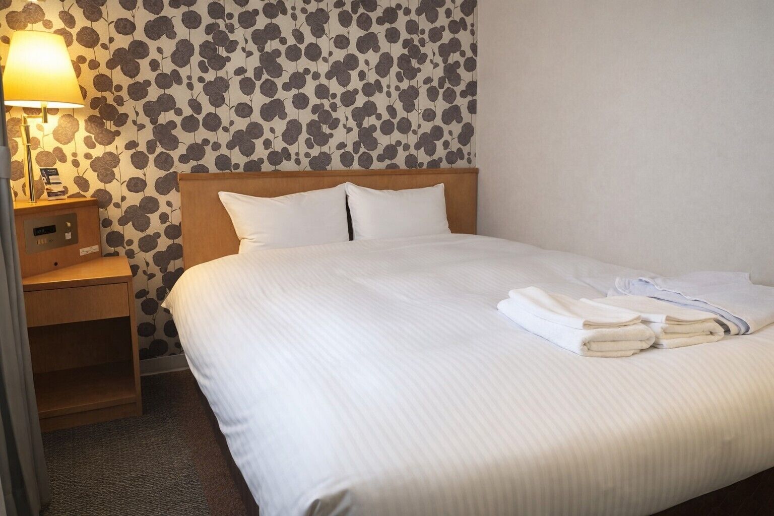 A neatly made bed with white linens and pillows, a bedside table with a lamp, and a wall with a floral pattern.