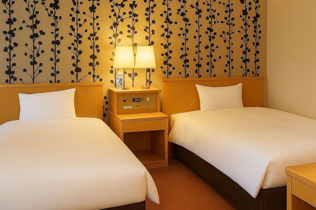 A hotel room with two beds, a nightstand with a lamp, and a wall with a floral pattern.