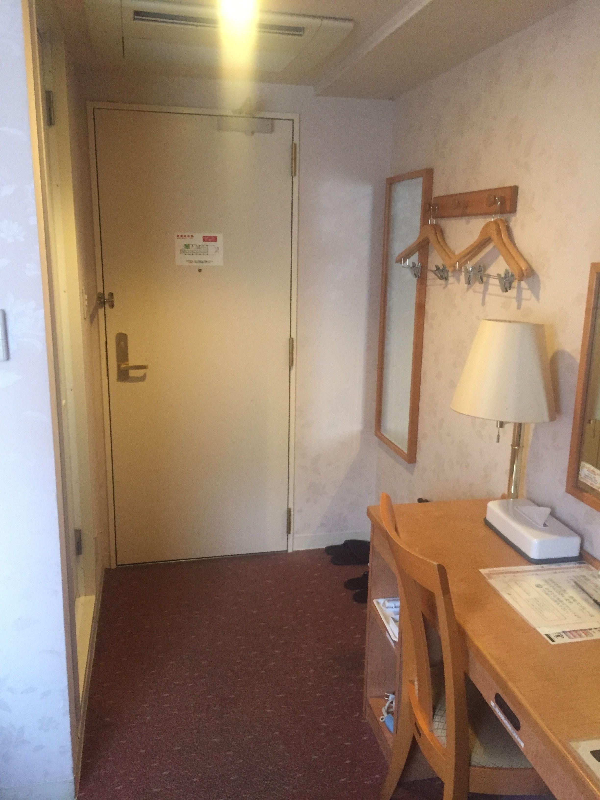 A hotel room with a closed door, a desk with a lamp, a chair, and a coat rack with hangers.