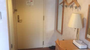 A hotel room with a closed door, a desk with a lamp, a chair, and a coat rack with hangers.