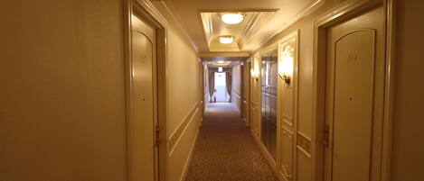 Corredor de hotel com pisos carpetados, paredes em tom creme e portas em ambos os lados.