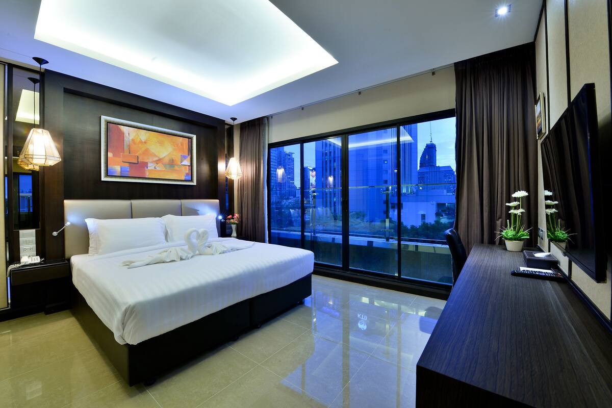 A modern hotel room with a large bed, a desk, and a city view through floor-to-ceiling windows.