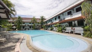 A swimming pool with lounge chairs and umbrellas, surrounded by a building with multiple balconies and palm trees. - Bahari Inn Tegal (Tegal)