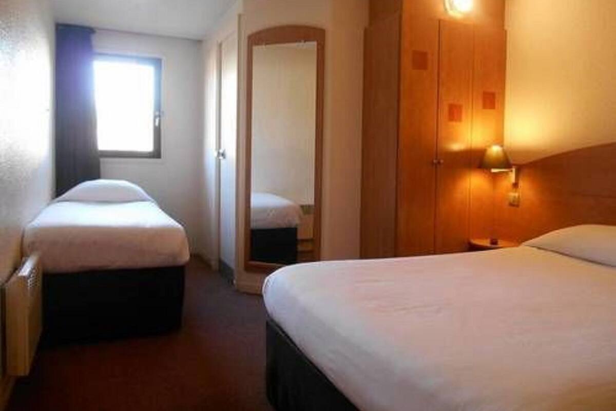 A hotel room with two single beds, a wooden wardrobe, a bedside table with a lamp, and a window with curtains.
