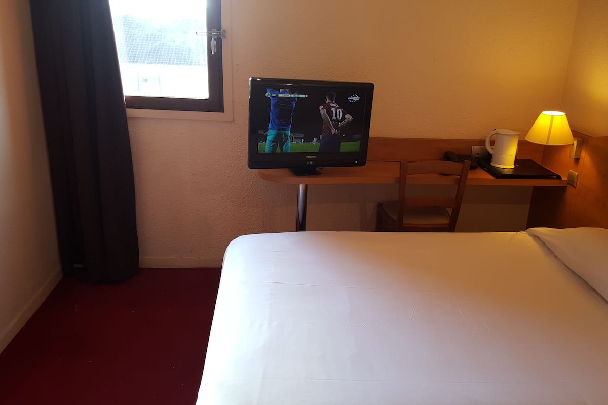 A hotel room with a bed, a desk with a computer monitor, a chair, a lamp, and a window.