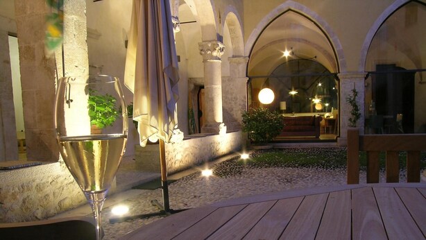 Une table en bois, un verre de vin blanc, un banc et un chemin de pierre menant à un espace extérieur aménagé avec vue sur l’intérieur d’un bâtiment.