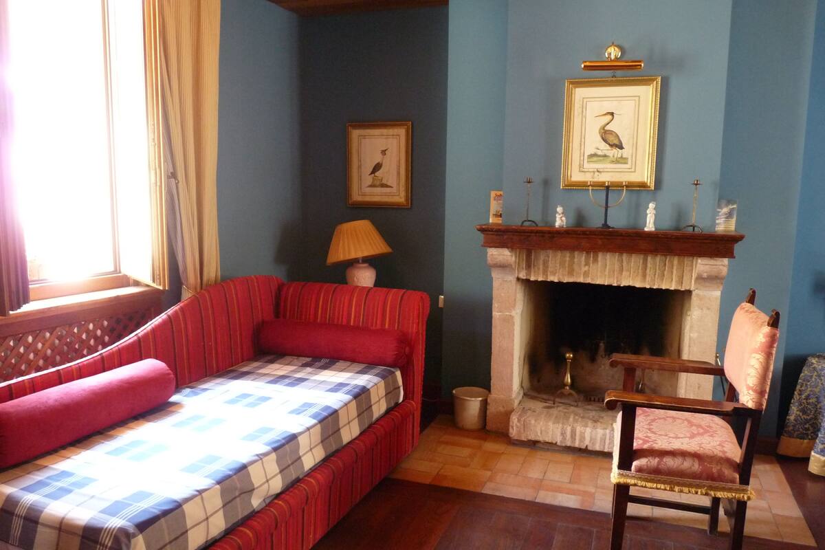 Ein Zimmer mit rotem Sofa, einer karierten Bettdecke, einem Kamin und einem darüber hängenden Bild, sowie einem Holzstuhl.