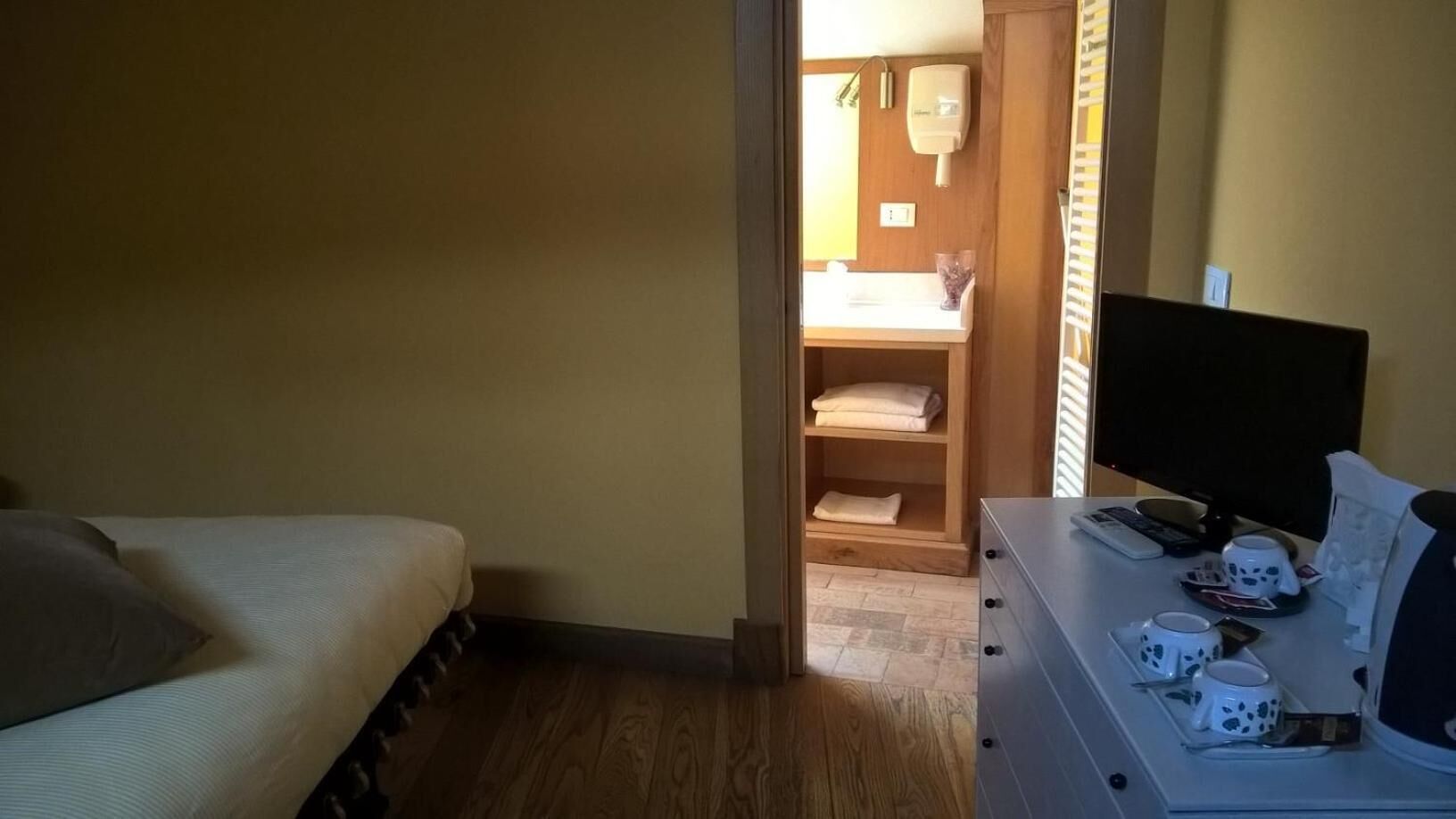A small room with a bed, a desk with a computer and mugs, and a wooden shelf with folded towels.