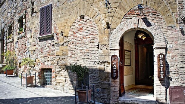 Un bâtiment en pierre avec une porte en bois portant l’inscription « BELVISTA » et « CAMER », ce qui indique une salle de contrôle vidéo. À l’extérieur, on trouve des plantes en pot et une petite table avec une chaise.
