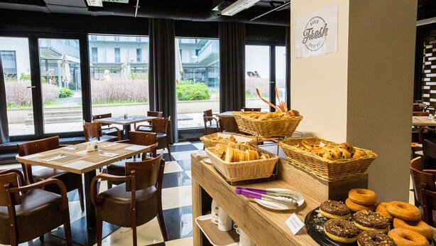 Una cafetería con suelo a cuadros, mesas y sillas de madera, cestas de pan y pasteles, y un letrero que dice 'Horneado fresco cada día'.