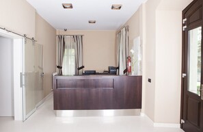 A reception area with a dark wood reception desk, a glass door, and a window with curtains. - Hotel Villa Marrón (Llanes)