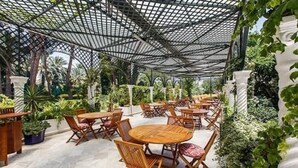 Een terras buiten met ronde houten tafels en stoelen, omgeven door weelderige begroeiing en witte zuilen.
