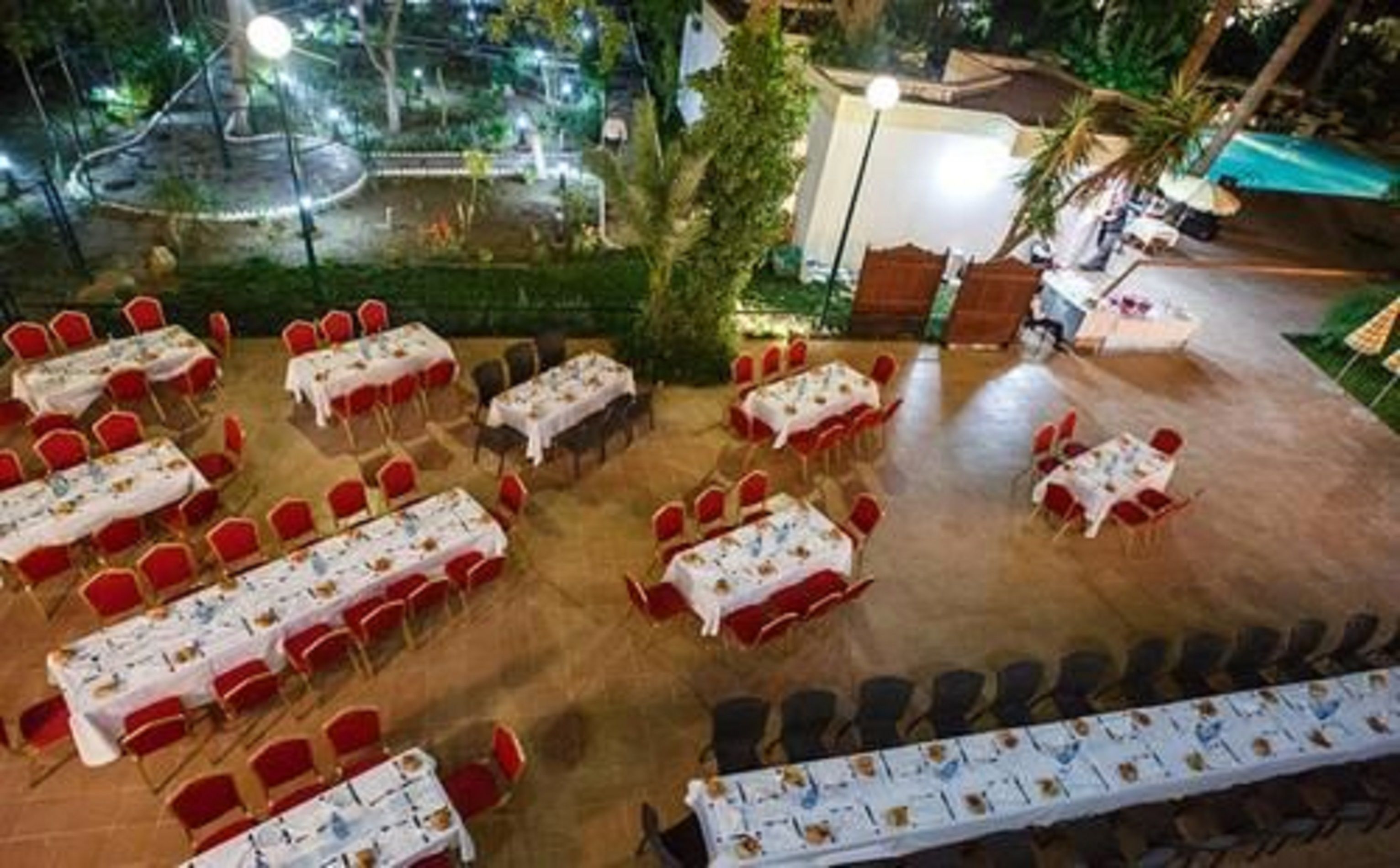 An outdoor dining area with multiple tables and chairs, surrounded by greenery and a pool.