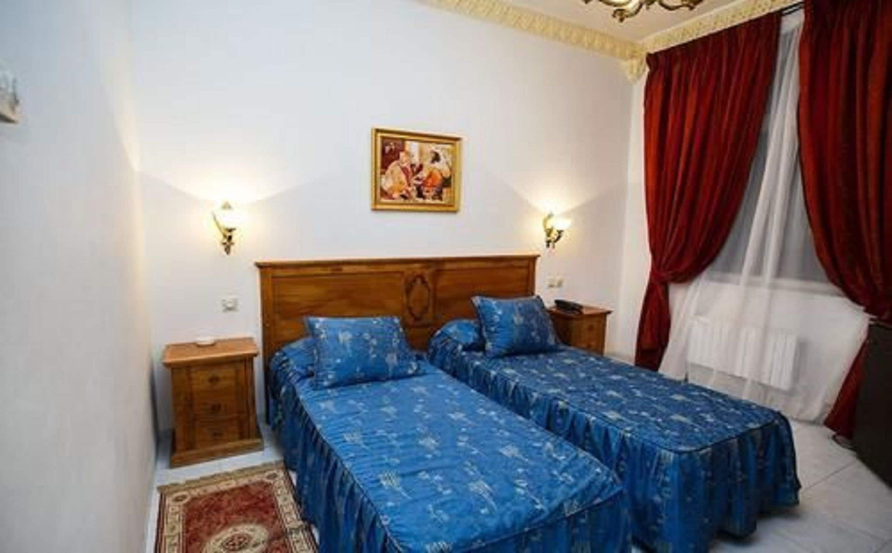 A bedroom with two beds, a wooden headboard, two bedside tables, a wall-mounted picture, and red curtains.