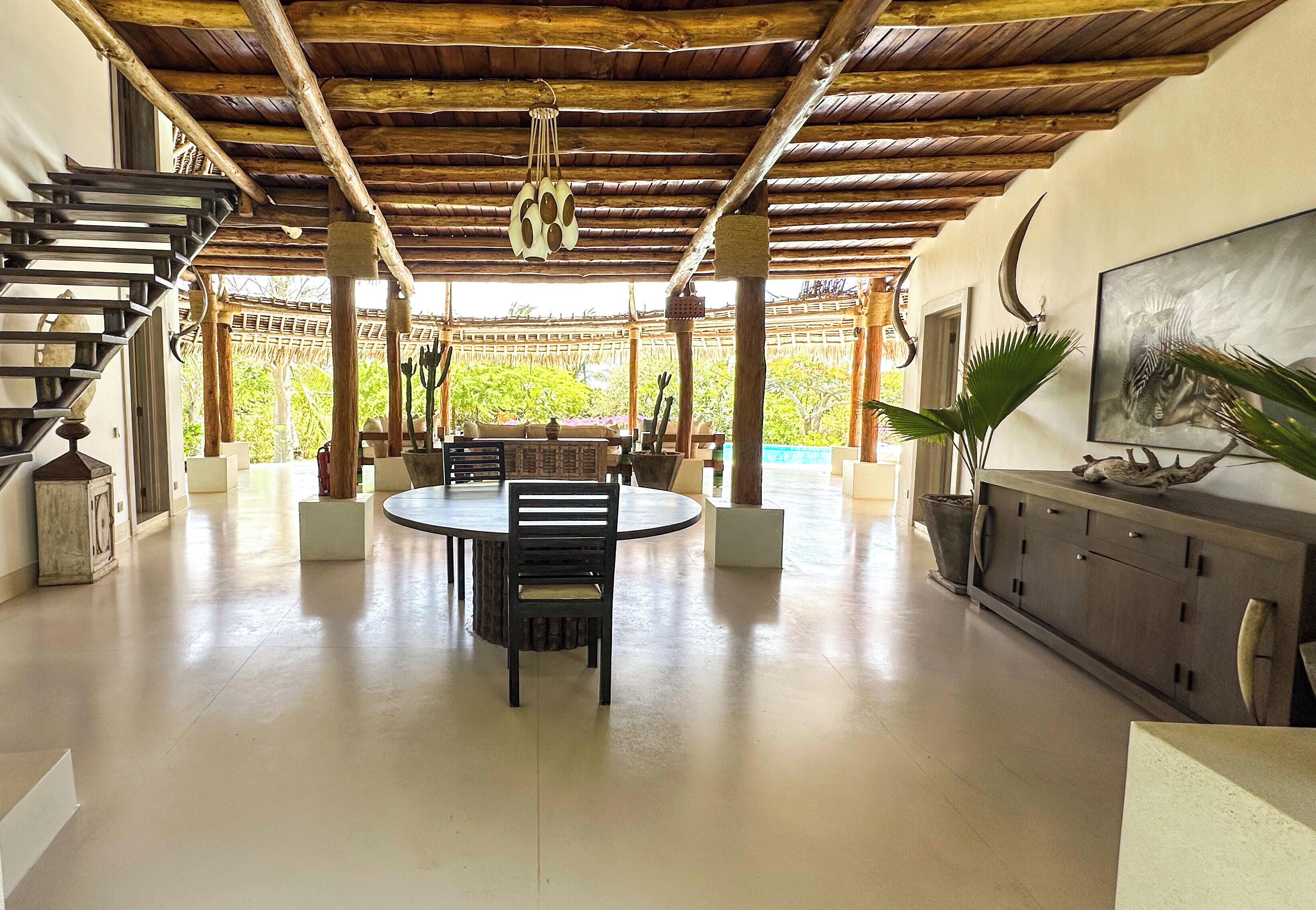 A spacious room with a wooden ceiling, a dining table with chairs, a sofa, a TV on a cabinet, and a staircase.