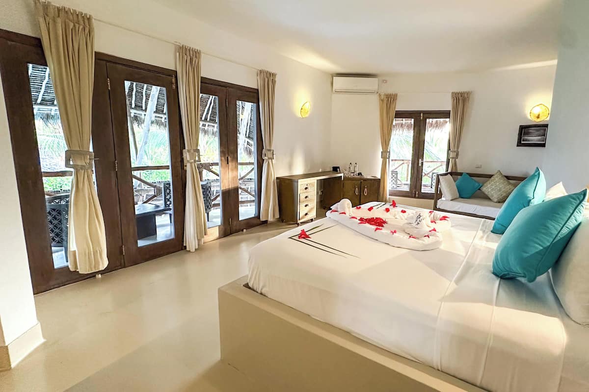 Quarto de hotel com cama, sofá, minibar e vista para o exterior através de grandes janelas.