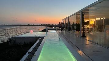 Un paseo marítimo con un edificio de vidrio, una fuente reflectante y vistas al atardecer.