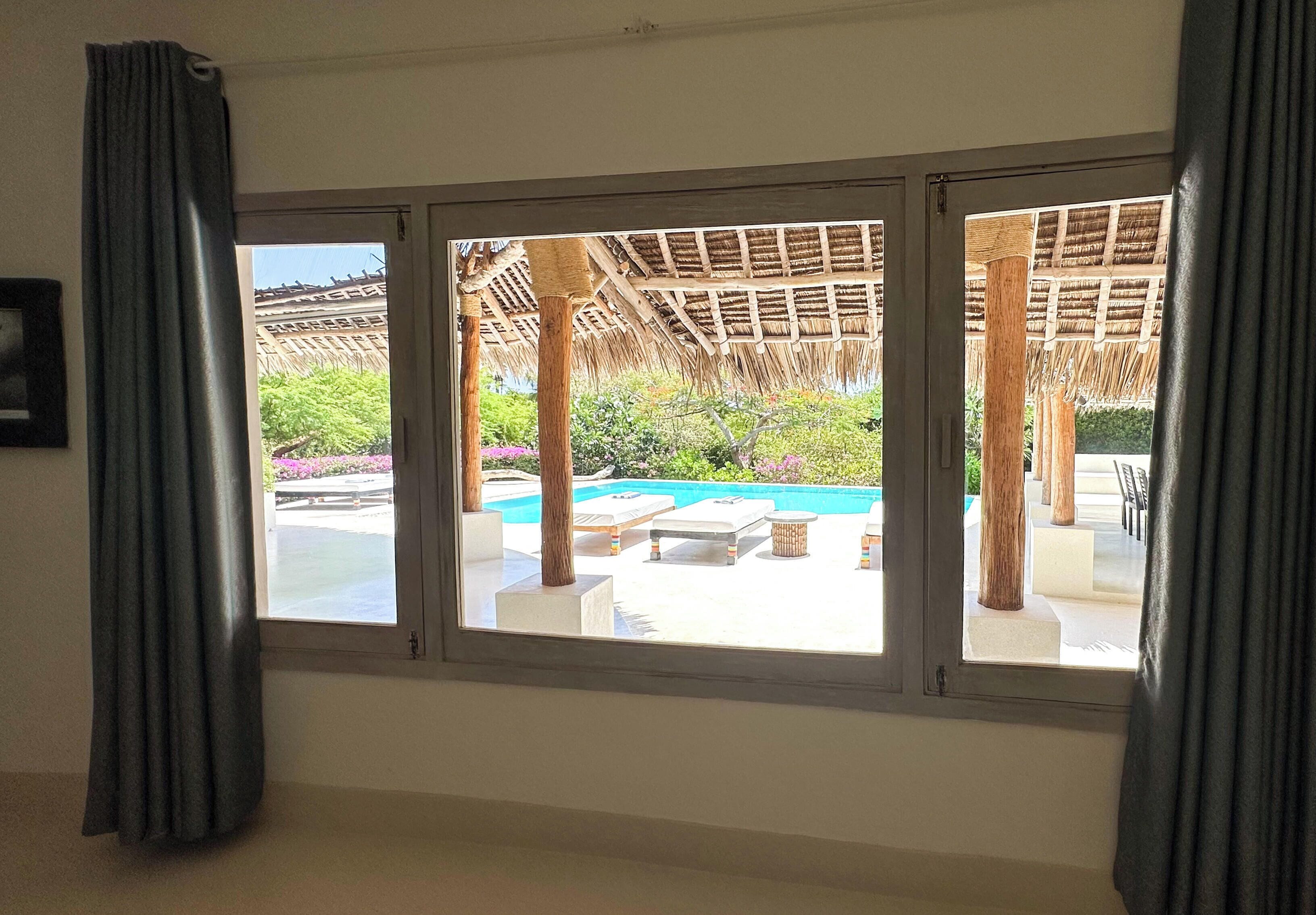 A room with a large window overlooking a pool area with lounge chairs and a thatched roof structure.
