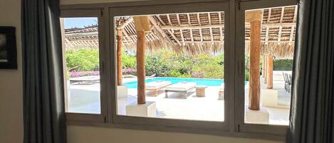 A room with a large window overlooking a pool area with lounge chairs and a thatched roof structure.