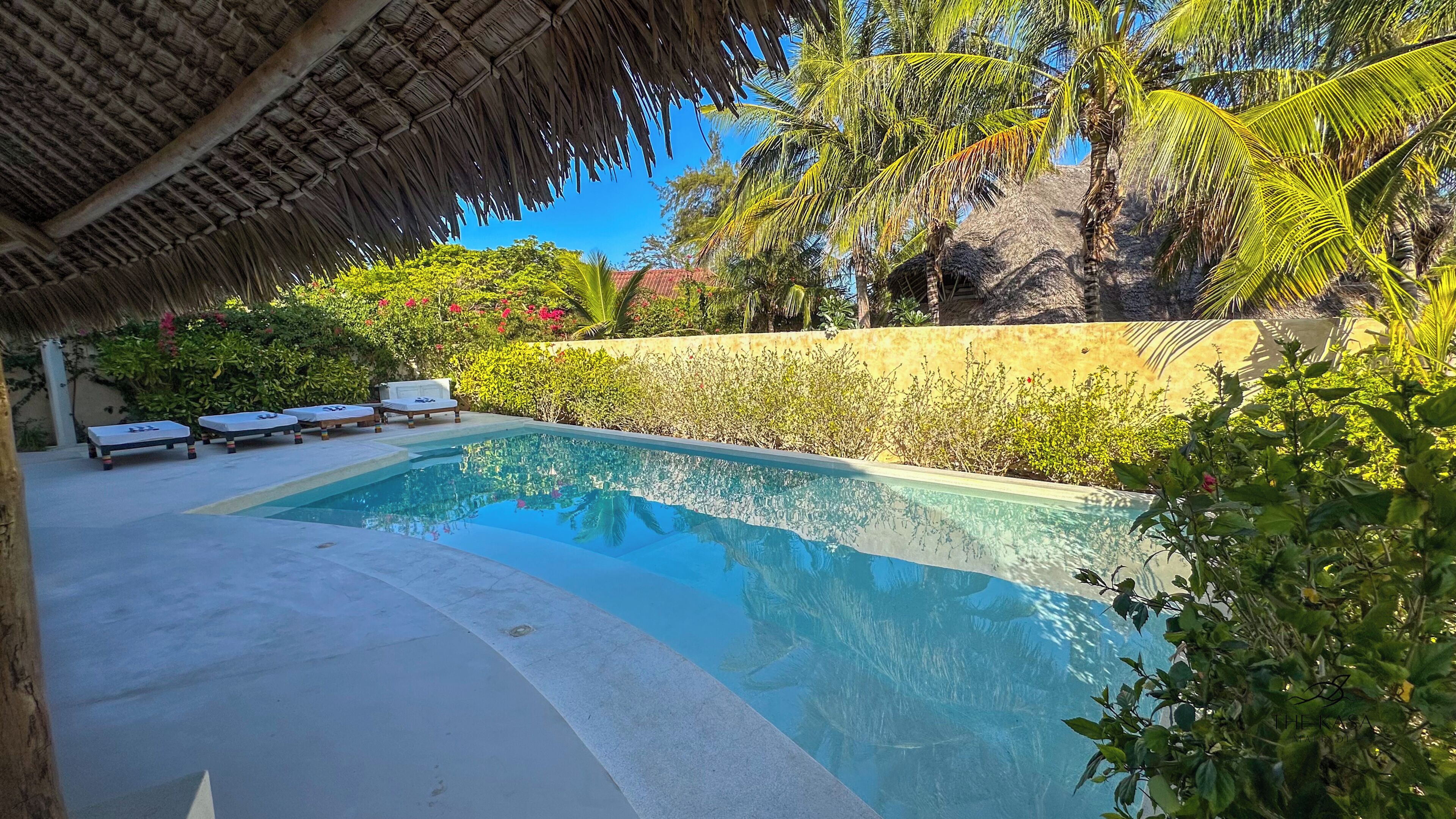 A swimming pool with lounge chairs, surrounded by lush greenery and a thatched roof structure.
