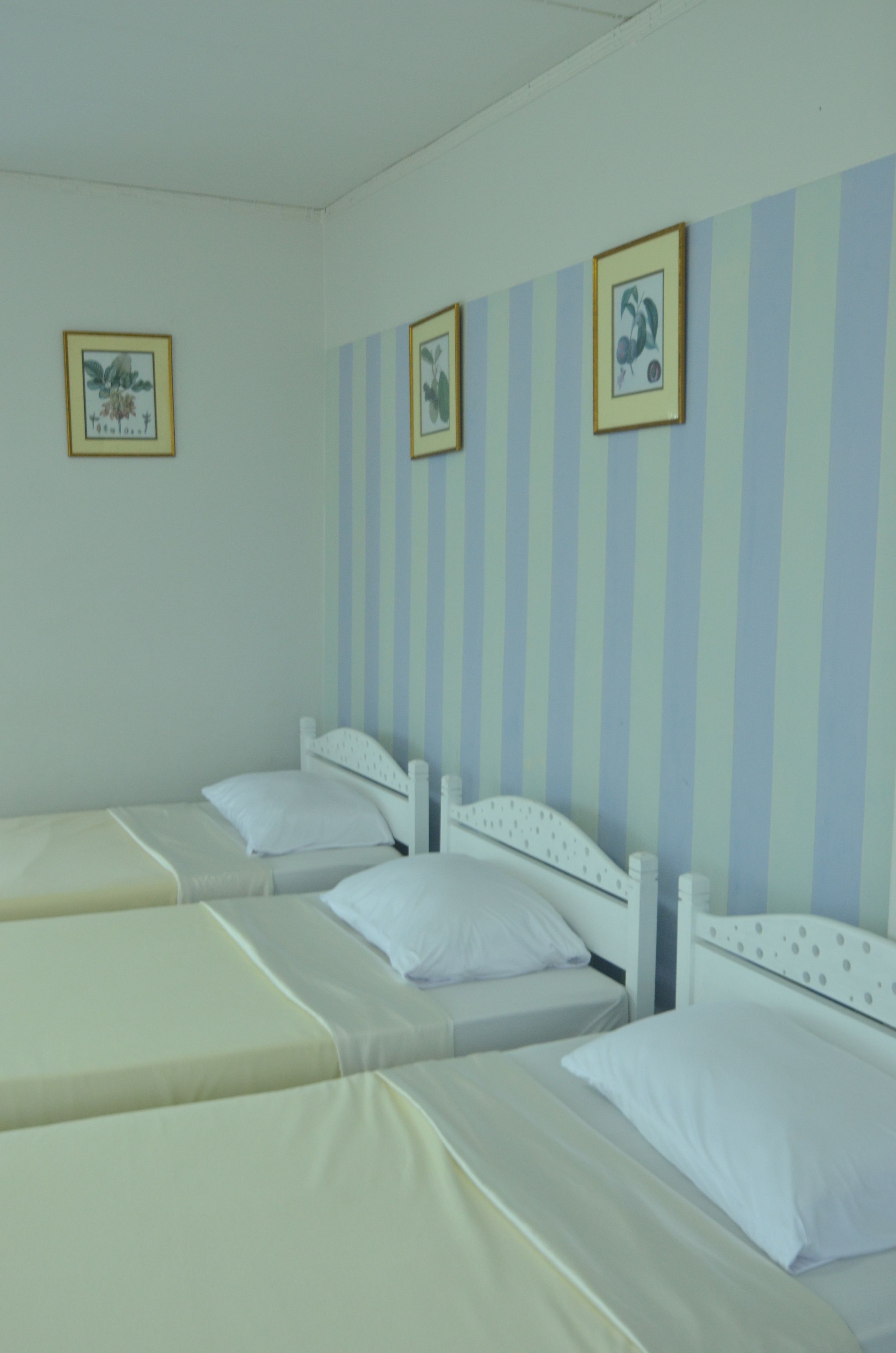 A room with three single beds, each with white bedding and headboards. The walls have light blue and white stripes, and there are framed pictures on the wall.