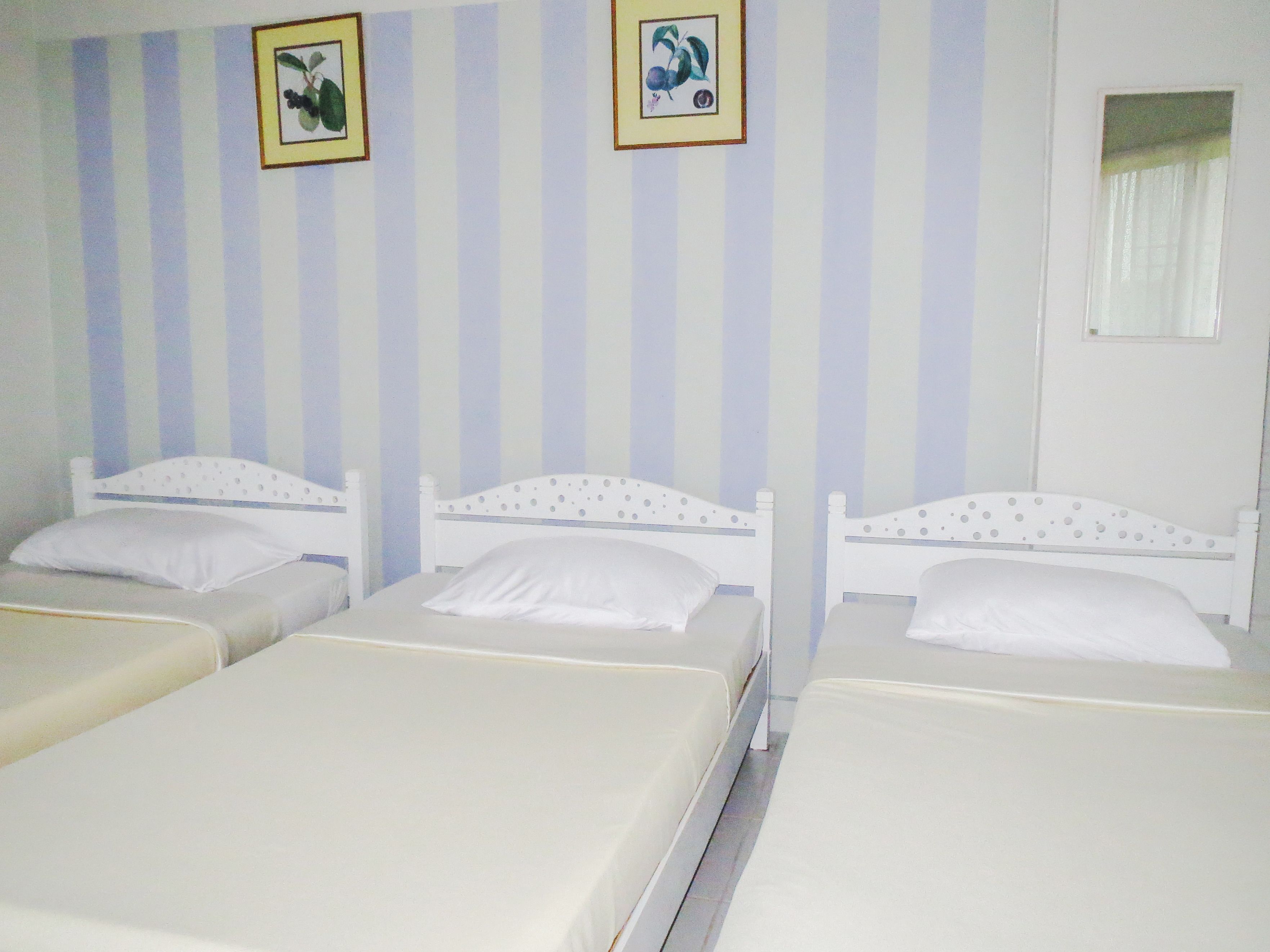 Een kamer met drie eenpersoonsbedden, elk met een wit hoofdbord en kussen. De muren hebben verticale strepen, en er hangen twee ingelijste foto’s aan de muur.