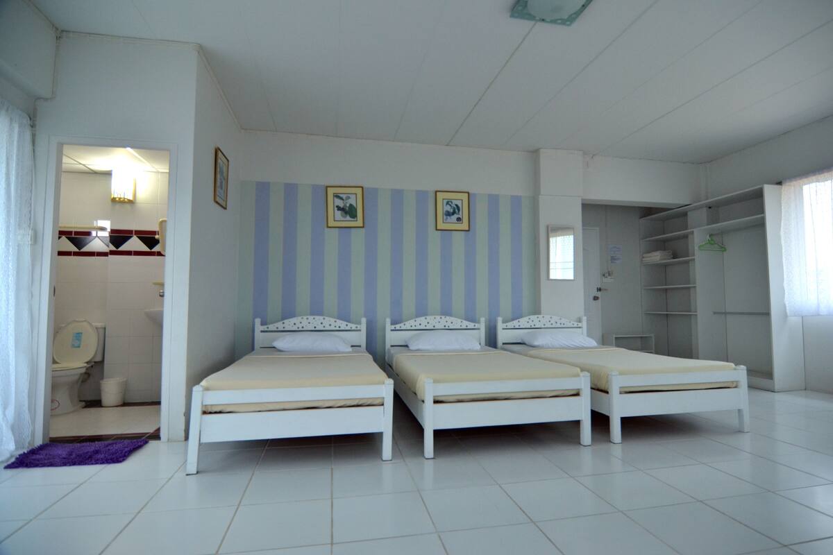 A room with three white beds, a bathroom with a toilet and sink, and a wardrobe with shelves.