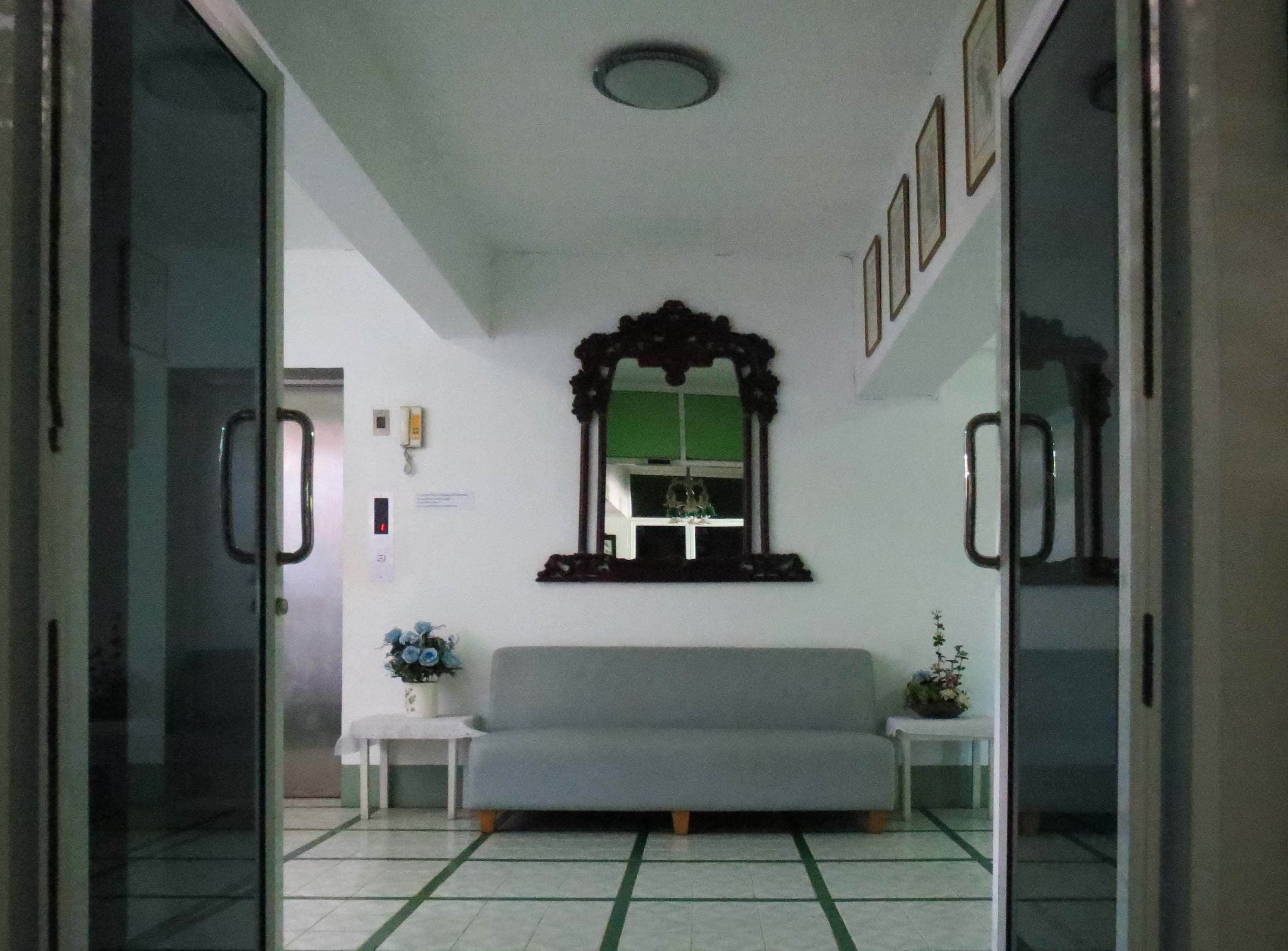 A room with a grey sofa, a small white table with a vase of flowers, a wall-mounted mirror, and a green-framed window.