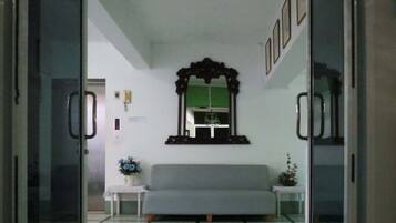 A room with a grey sofa, a small white table with a vase of flowers, a wall-mounted mirror, and a green-framed window.