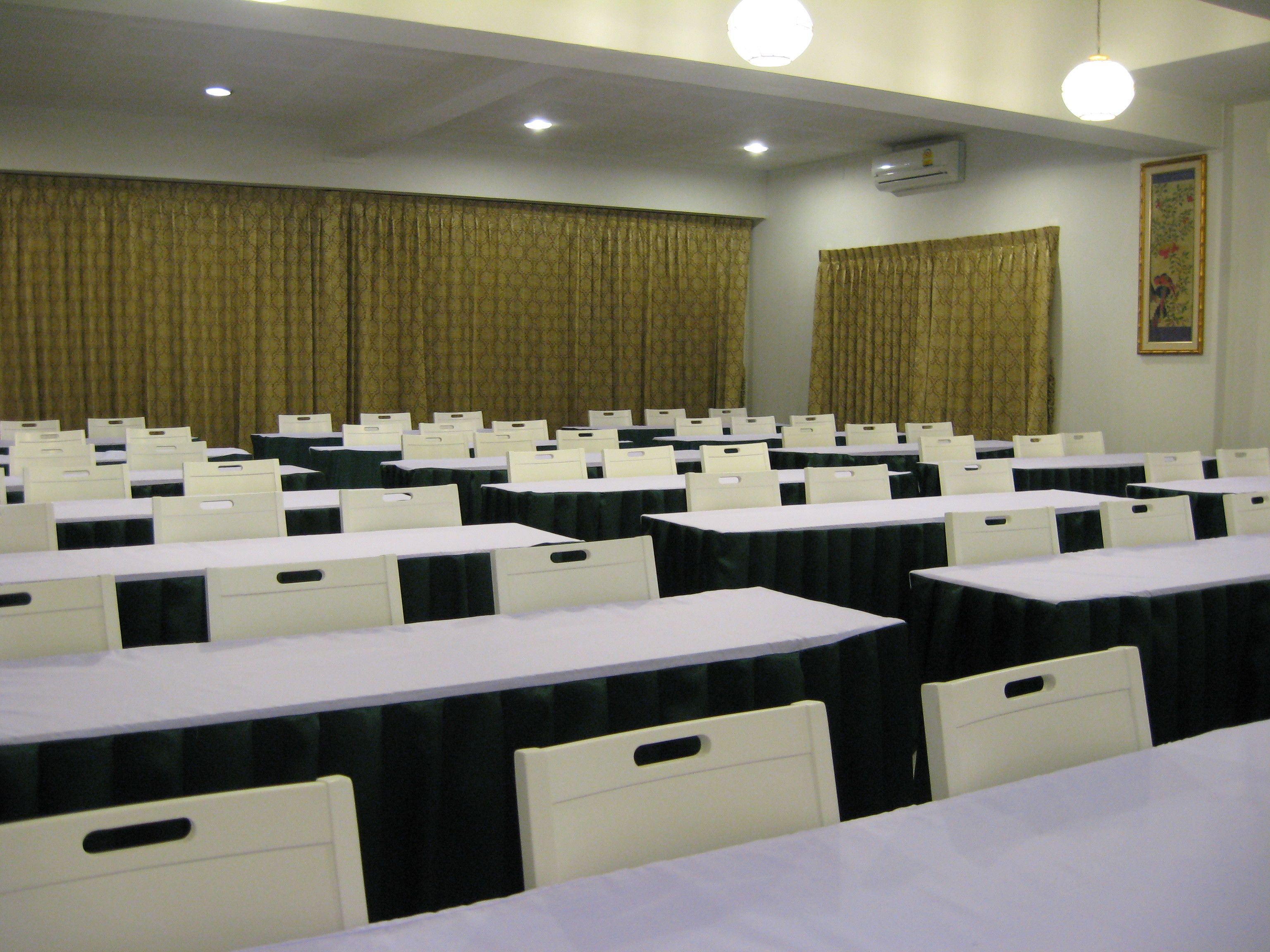 Une salle de conférence avec des rangées de tables et de chaises, un plafond doté d’un éclairage encastré et des rideaux aux fenêtres.
