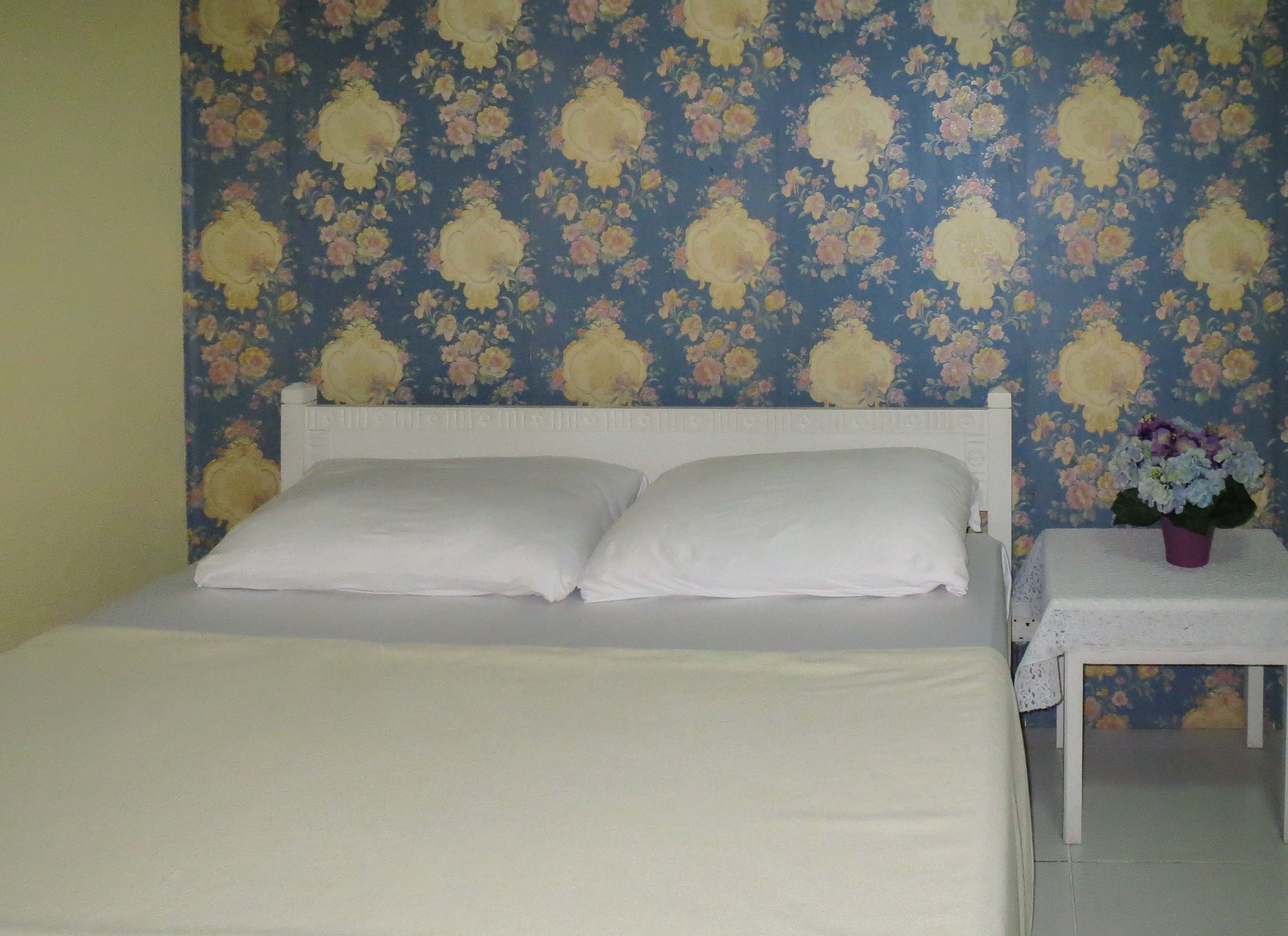 Ein Bett mit weißer Bettwäsche, floraler Tapete und einem kleinen Tisch mit Blumenvase.