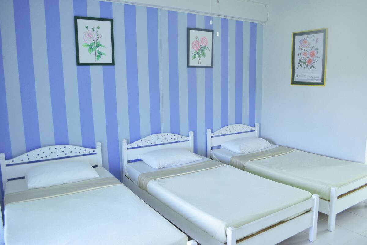A room with three single beds, each with a white headboard and mattress. The walls have a blue and white striped pattern, and there are framed floral pictures on the wall.