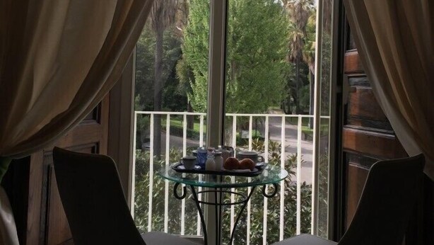 Un balcón con una mesa de vidrio y dos sillas, con vista a una calle con árboles y edificios.