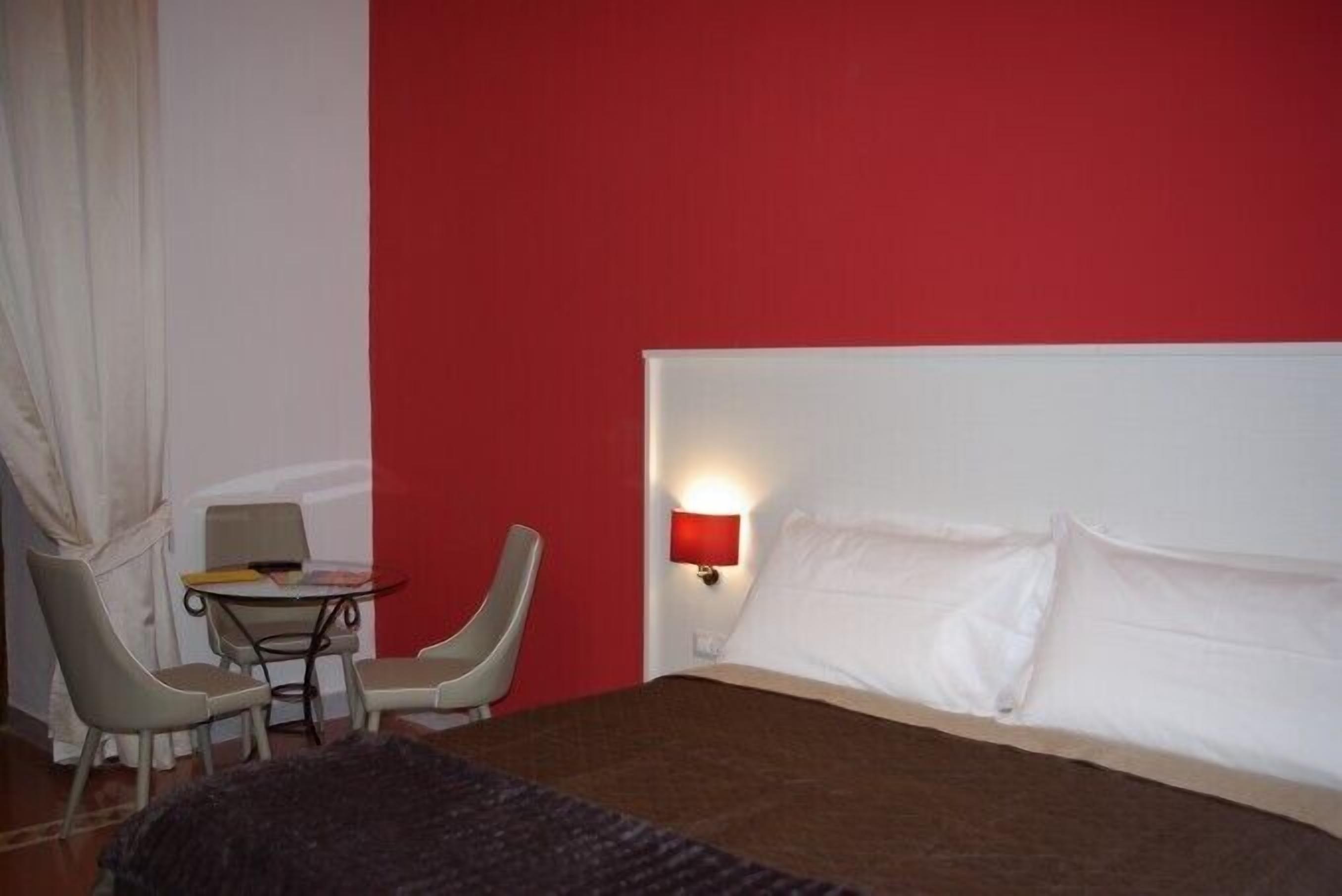 A hotel room with a bed, a small table with chairs, a red wall, and a nightstand with a lamp.
