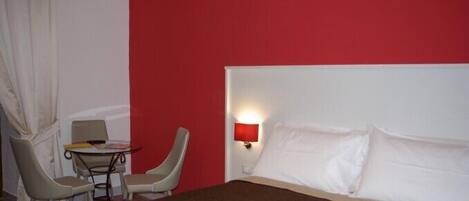 A hotel room with a bed, a small table with chairs, a red wall, and a nightstand with a lamp.